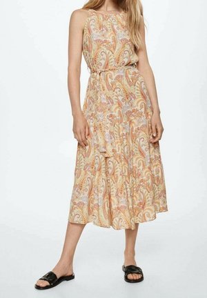 Femme portant une robe midi sans manches avec imprimé cachemire beige et orange et des sandales plates noires, debout sur fond blanc.