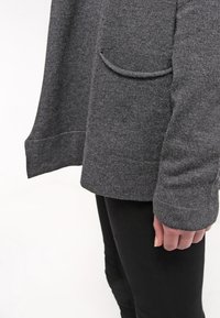 Cardigan gris de punto con un corte holgado, que incluye un bolsillo único y puños acanalados, combinado con pantalones negros. Textura suave, diseño casual.