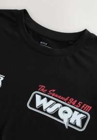 Czarny bawełniany t-shirt z okrągłym dekoltem, z etykietą "NETFLIX" oraz nadrukowanymi grafikami: "The Squawk 94.5 FM" i "WSQK" w kolorze białym i różowym.