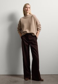 Giovane donna con capelli biondi che indossa un maglione beige oversize e pantaloni larghi marrone scuro, in piedi contro un muro bianco con le mani nelle tasche.