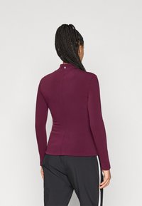 Μπλούζα με μακριά μανίκια σε βαθύ βουργουνδί ύφασμα με ψηλό γιακά. Διαθέτει μικρό λογότυπο στο πίσω μέρος του λαιμού· ομαλή υφή, εφαρμοστή σχεδίαση.