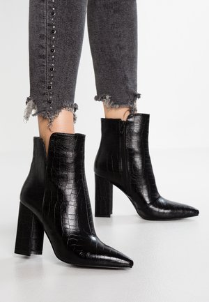 Botas de tobillo negras de punta afilada con textura de cocodrilo y tacones de bloque, combinadas con jeans oscuros de dobladillo desgastado que presentan studs en negro y plata.