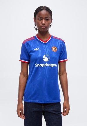 Persona giovane che indossa la maglia da calcio blu del Manchester United con i loghi di Snapdragon e Adidas, in piedi contro uno sfondo bianco uniforme.