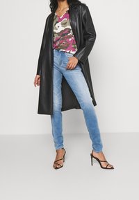 Svart konstläder långkappa över en kamouflage V-ringad skjorta i rosa, grönt och beige, i kombination med ljusblå jeans och svarta remsandaler.