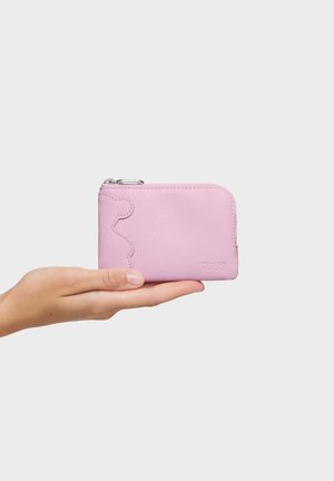 Mano sosteniendo una pequeña cartera rosa con cremallera, superficie texturizada y contorno de oso en relieve en el frente, sobre un fondo liso.