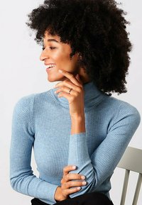 Lyseblå ribbestrikket turtleneck sweater med lange ærmer. Har en tætsiddende pasform og struktureret stof, som komplementerer en siddende model med krøllet hår.