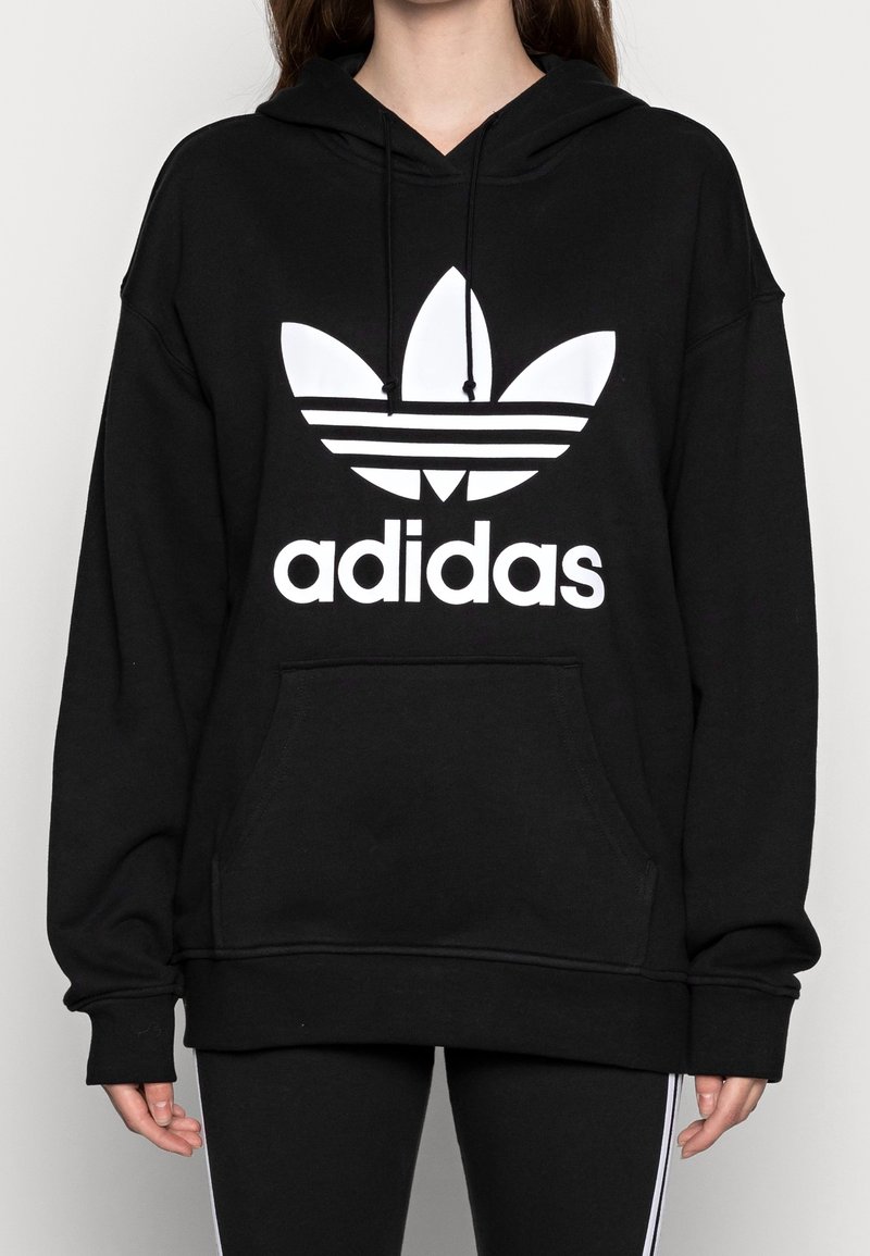 Černá mikina s kapucí vyrobená z měkké látky, s velkým bílým logem Adidas a přední kapsou na klokaní styl. Uvolněný střih s dlouhými rukávy.