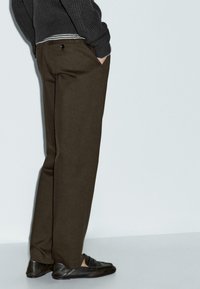 Pantaloni marroni su misura con una texture liscia, caratterizzati da una tasca posteriore con un bottone nero, abbinati a mocassini neri e a un maglione grigio.