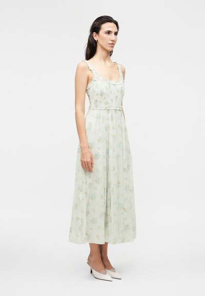 Γυναίκα με μακριά σκούρα μαλλιά που φοράει αμάνικο, ελαφρύ μεσαίο φόρεμα με λουλούδια και άσπρα μυτερά τακούνια, στέκεται μπροστά σε μονόχρωμο φόντο.