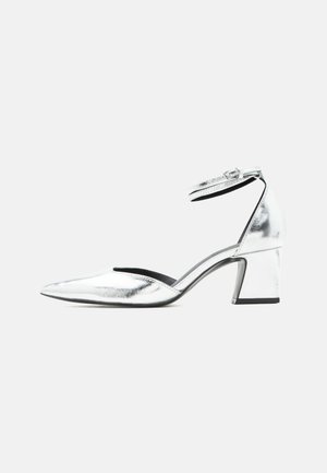Silberfarbener Metallic-Schuh mit niedrigem Blockabsatz, spitzer Toe und Knöchelriemen mit Schnalle auf weißem Hintergrund.