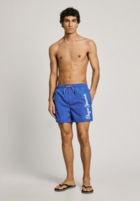 Μπλε μαγιό με κορδόνι στη μέση, διακοσμημένο με λευκό γραφικό κείμενο στο πλάι, ελαφρύ ύφασμα και λεία υφή.