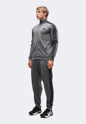 Joven de pie, vestido con un chándal gris con rayas negras y zapatillas deportivas en blanco y negro, mirando ligeramente hacia la izquierda.