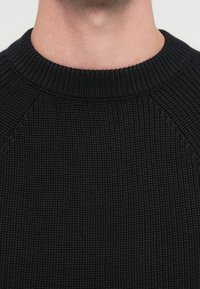 Pulover negru tricotat, cu textură cu dungi, decolteu rotund și mâneci raglan. Materialul pare gros și călduros.