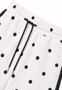 Tissu blanc à pois noirs, taille élastique et cordons noirs avec embouts métalliques, montrant une vue partielle du pantalon.