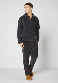 Ciemnoszara kurtka sztruksowa z kołnierzem i dwoma przednimi kieszeniami, zestawiona z pasującymi spodniami i brązowymi zamszowymi butami. Gładka faktura.