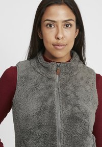 Grau Fleece-Weste mit einem hohen Kragen und einem Reißverschluss vorne. Tragen über einem bordeauxfarbenen Langarmtop, das eine weiche, strukturierte Oberfläche aufweist.