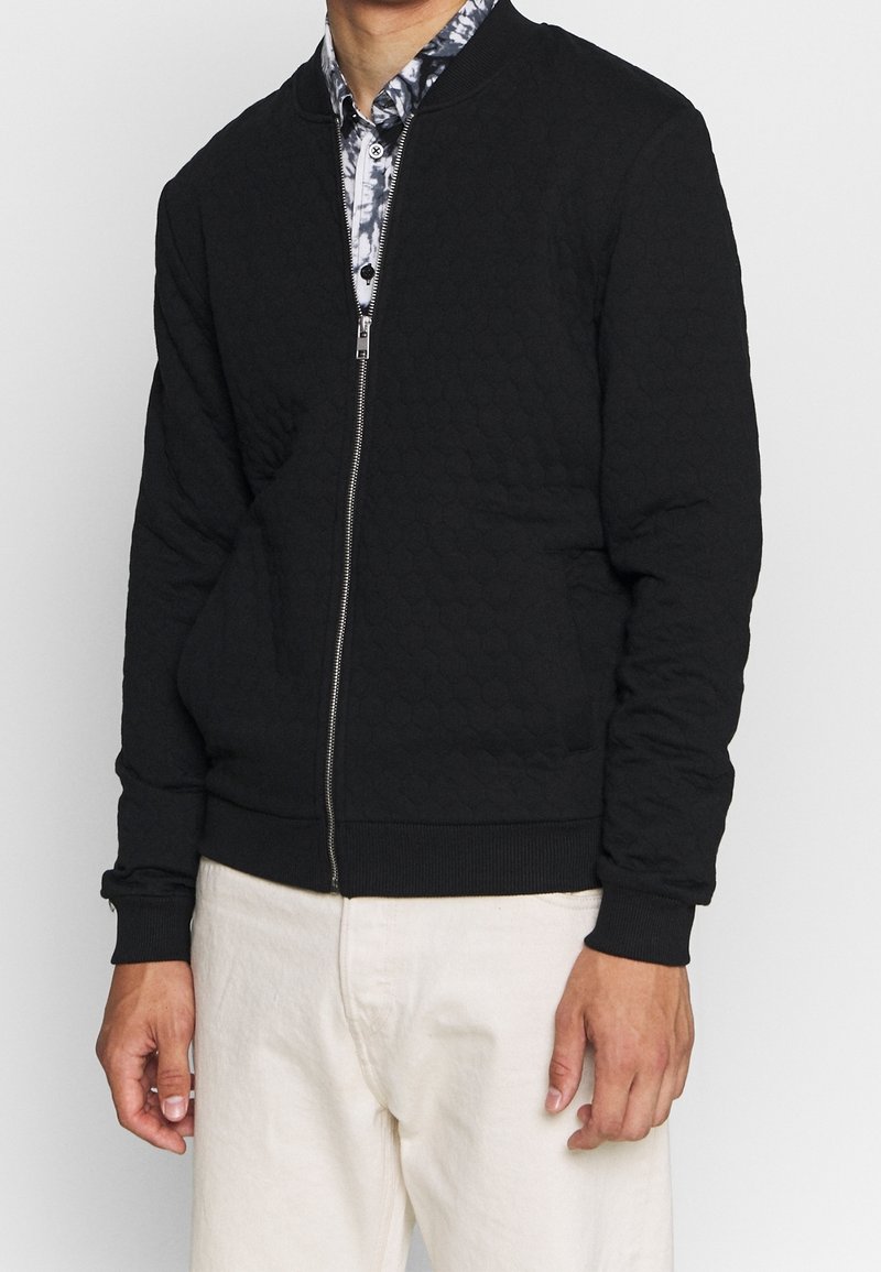 Homme portant une veste matelassée noire zippée par-dessus une chemise à motifs noirs et blancs, avec un pantalon écru, sur un fond uni.