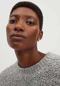 Maglione a maglia grigio chiaro, texturizzato, con scollatura rotonda, realizzato in un materiale morbido e soffice. Orecchino a cerchio argentato visibile nell'orecchio sinistro.