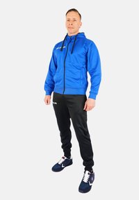 Sudadera azul con cremallera, pantalones de chándal negros y zapatillas de deporte azul marino. La sudadera tiene cordones y un logotipo, y los pantalones cuentan con un detalle de marca en blanco.