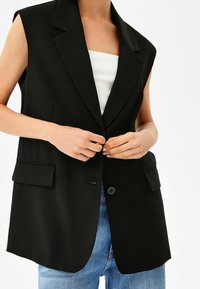 Svart ärmlös blazer med kantslag, knappstängning fram och två fickor. Bärs över en vit ribbad linne och denimjeans.