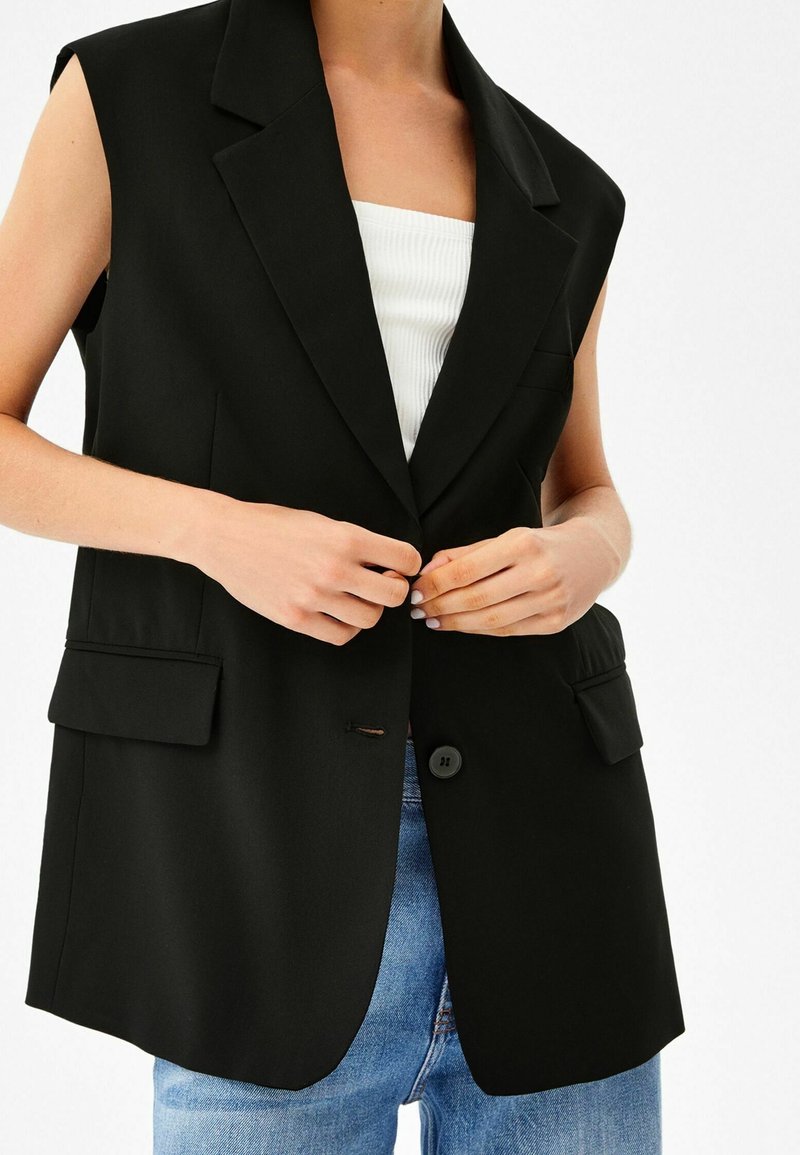 Svart ärmlös blazer med kantslag, knappstängning fram och två fickor. Bärs över en vit ribbad linne och denimjeans.