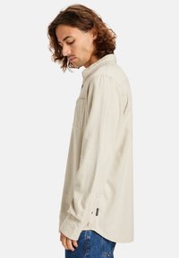 Beige long-sleeve shirt with a collar, a front pocket, and side splits. Soft texture, relaxed fit, paired with blue jeans.