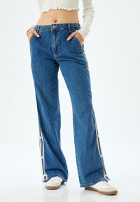 Pantalones de mezclilla azul con un corte holgado, cintura alta, botones laterales y detalles a rayas en blanco y negro a lo largo de las costuras exteriores.
