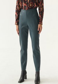 Pantaloni in pelle teal con vita alta, taglio slim e design senza cuciture. Indossati con stivali neri con tacco alla caviglia.