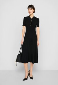 Svart ribbad midiklänning med korta ärmar, rund halsringning, gulddetaljerade knappar och veckad kjol. Bärs med svarta spetsiga klackar och en handväska.