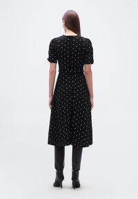 Γυναίκα με μακριά καστανά μαλλιά, φορώντας μαύρο μίντι φόρεμα με λευκές πουά, φουσκωτά μανίκια και μαύρες ψηλοτάκουνες μπότες μέχρι το γόνατο, με την πλάτη γυρισμένη.