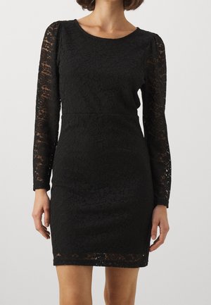 Vrouw die een zwarte, nauwsluitende kanten jurk met lange mouwen en bloemmotieven draagt, staand tegen een effen lichte achtergrond.