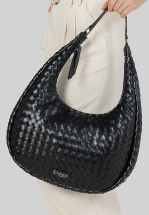 Main tenant un grand sac porte-épaule en cuir tissé noir avec une sangle courte, porté sur un pantalon de couleur crème.