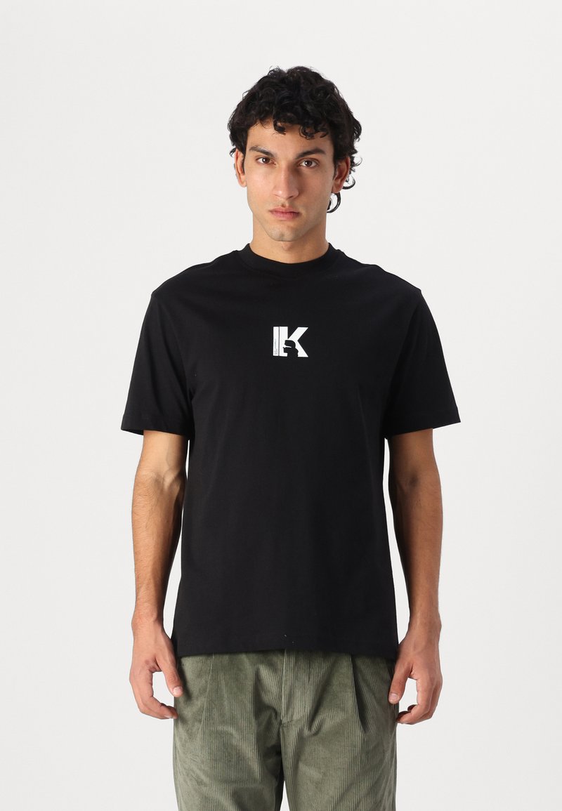 Zwarte katoenen T-shirt met ronde hals, voorzien van een witte graphic logo in het midden van de borst. Model draagt olijfgroene corduroy broek.