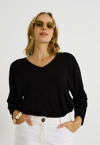 Top negro de manga larga con escote en V, tejido suave y corte relajado. Combinado con pantalones blancos de talle alto y una pulsera de brazalete dorada. Gafas de sol con lente tintada en verde.