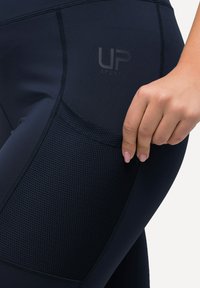 Leggings da allenamento blu navy con una tasca in rete, caratterizzati da una superficie liscia, cuciture a contrasto e un logo impresso sul lato.
