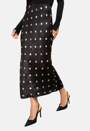 Schwarzer Rock mit goldenen Punkten, aus einem glatten, strukturierten Material. Hat ein langes, gerades Design und einen Seitenslitz. Kombiniert mit schwarzen High Heels.