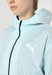 Lekka jasnoniebieska kurtka z kapturem, z czarnym zamkiem błyskawicznym i refleksyjnym białym logo Puma na lewej piersi. Gładka tekstura.