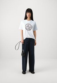 T-shirt bleu clair à manches courtes avec un design graphique circulaire marron. Associé à un pantalon large marine et un petit sac à bandoulière noir.