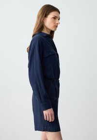 Vestito camicetta blu navy realizzato in tessuto testurizzato, con colletto, chiusura anteriore con bottoni, due tasche sul petto, maniche lunghe e vita con cintura.