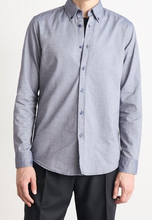 Homme portant une chemise bleu clair à manches longues avec boutons et un petit logo "BOSS" sur la poitrine, associée à un pantalon noir.