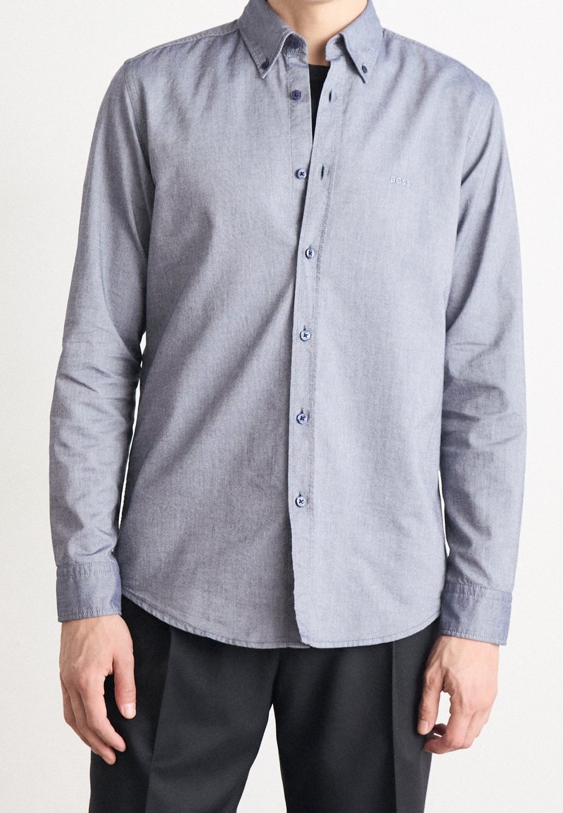 Homme portant une chemise bleu clair à manches longues avec boutons et un petit logo "BOSS" sur la poitrine, associée à un pantalon noir.