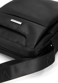 Borsa a tracolla nera in tessuto con superficie texturizzata, tasca frontale, placca con logo in argento e una larga tracolla regolabile con branding.