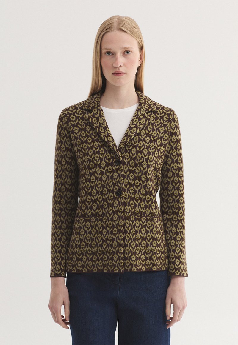Femme aux cheveux blonds et raides portant un blazer marron et jaune à motif floral sur une chemise blanche et un pantalon bleu foncé, debout devant un fond uni.