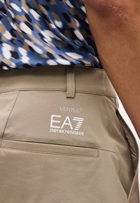 Μπεζ σορτς Emporio Armani EA7 Ventus σε άτομο που φορά μπλε πουκάμισο με σχέδια, με προβολή της πίσω τσέπης και της λεπτομέρειας του λογότυπου.