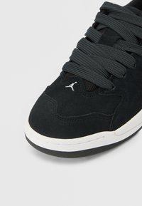 Μύτη μαύρου σουέντ αθλητικού παπουτσιού με παχιά μαύρα κορδόνια, λευκή μέση σόλα και μικρό λευκό λογότυπο Jumpman πάνω κοντά στα κορδόνια.