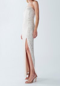 Abito aderente senza spalline in beige metallico con paillettes; presenta una spacco alto fino alla coscia su un lato; fodera interna liscia; indossato con tacchi nudi.