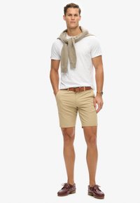 Pantalones cortos beige, camiseta blanca y un suéter marrón claro sobre los hombros. Cinturón marrón, zapatos rojo oscuro con cordones. Diseño simple, estilo casual.