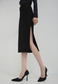 Jupe midi noire ajustée avec une fente sur le côté, associée à des escarpins noirs à bout pointu. Tissu lisse, design simple et lignes épurées.