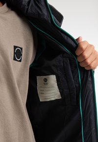 Zwarte jas met een teal rits, voorzien van een gladde binnenvoering en een beige shirt met een klein zwart logo op de borst.