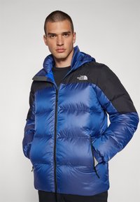 Niebieska i czarna puchowa kurtka z zamkiem z przodu, kapturem i bocznymi kieszeniami, wykonana z błyszczącego, wodoodpornego materiału, z logo na górnej lewej stronie.